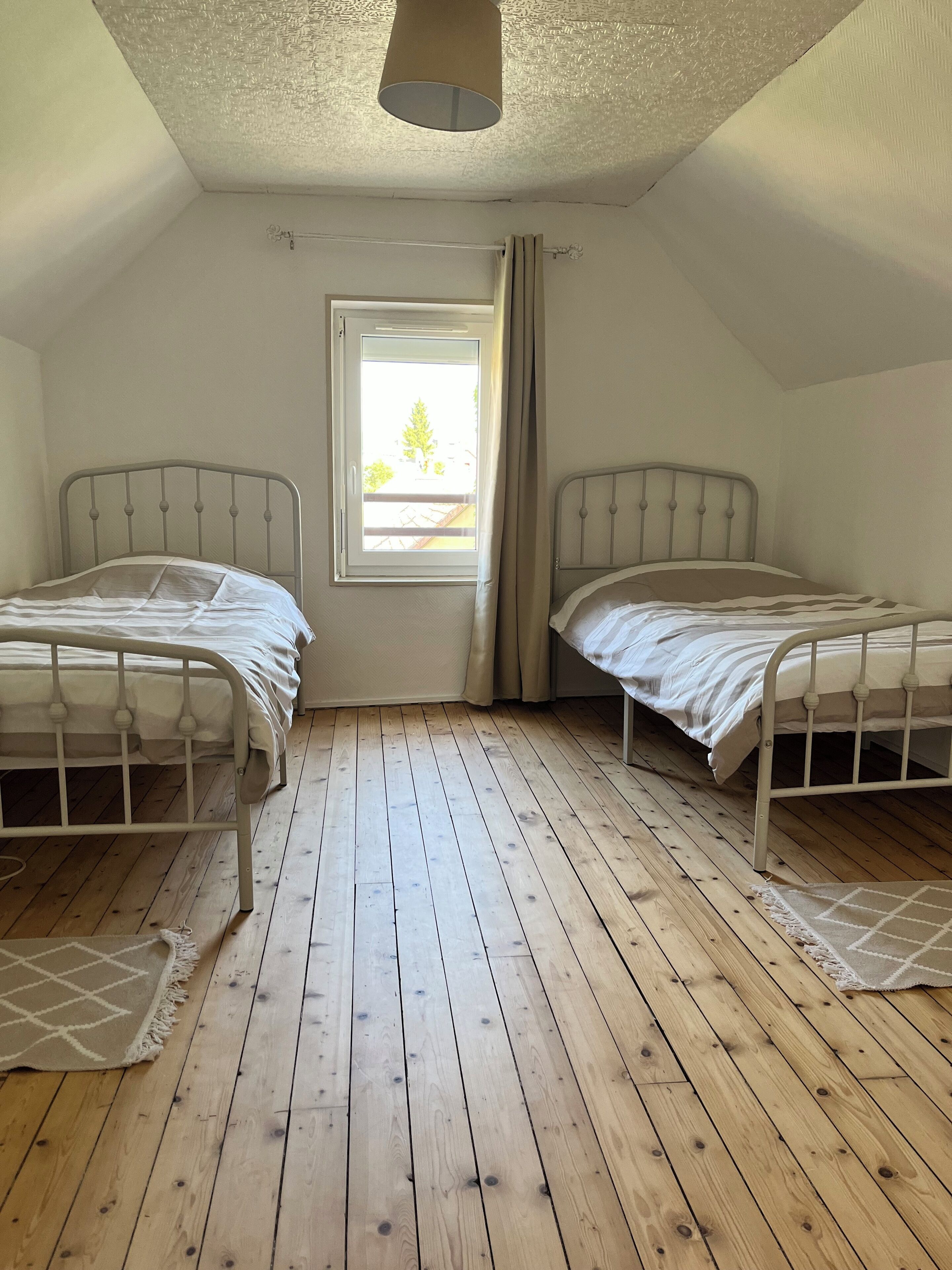 Eine kleine Einzimmerwohnung mit zwei Etagenbetten aus Metall, Holzboden, einem Fenster mit Vorhängen und einer Pendelleuchte.