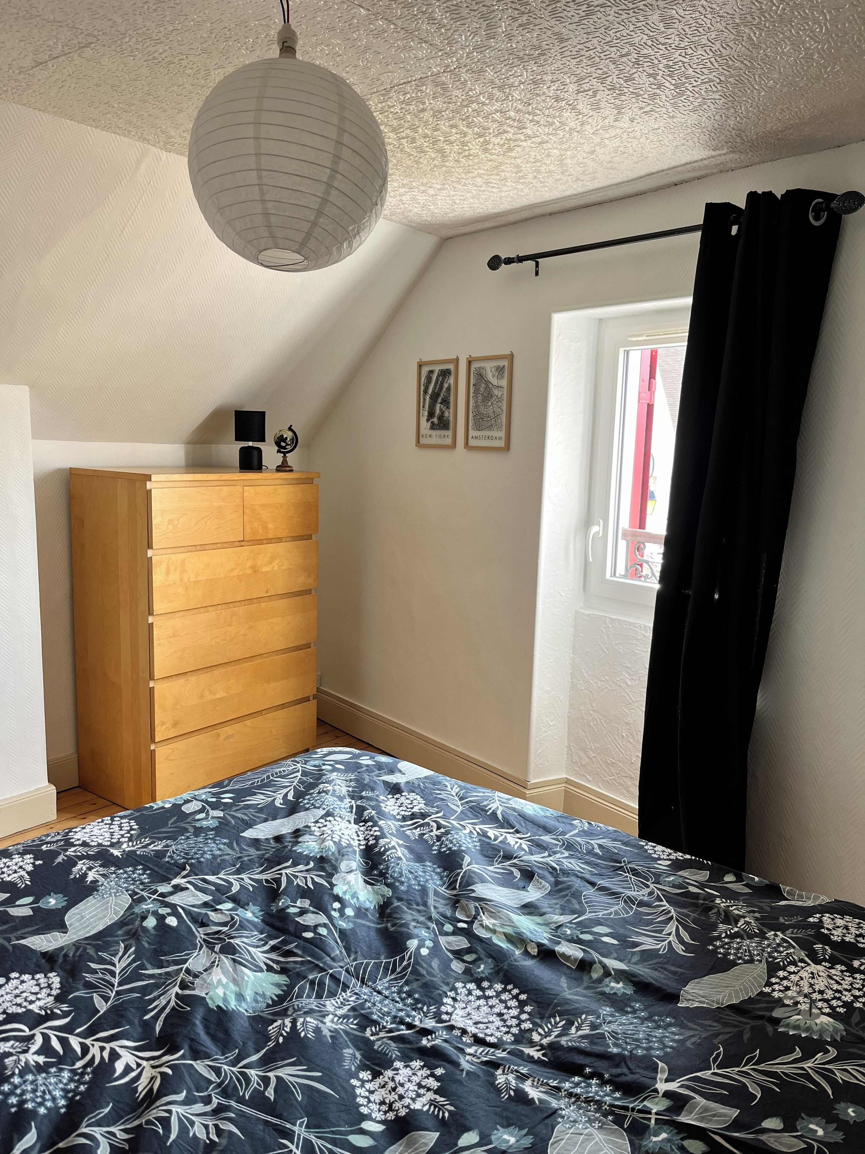 Un dormitorio con cama, cómoda de madera, lámpara colgante y dos mapas en la pared.