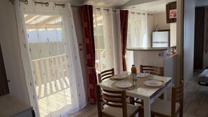 Comedor con mesa dispuesta para cuatro sillas, una pequeña cocina con refrigerador y vista a través de una ventana con cortinas.