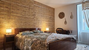 Une chambre à coucher avec une tête de lit en bois, un lit recouvert d’une literie à motifs, des lampes de chevet, un bureau et une fenêtre avec des rideaux.