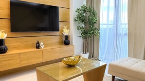 Ein modernes Wohnzimmer mit einem Flachbildfernseher an einer Holzwand, einem Glastisch, einem beigefarbenen Sofa und einem großen Fenster mit Vorhängen.