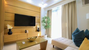 Ein modernes Wohnzimmer mit einem flachbildfernseher an einer Holzwand, einem Glastisch, einem beigefarbenen Sofa mit blauen Kissen und einer Topfpflanze.