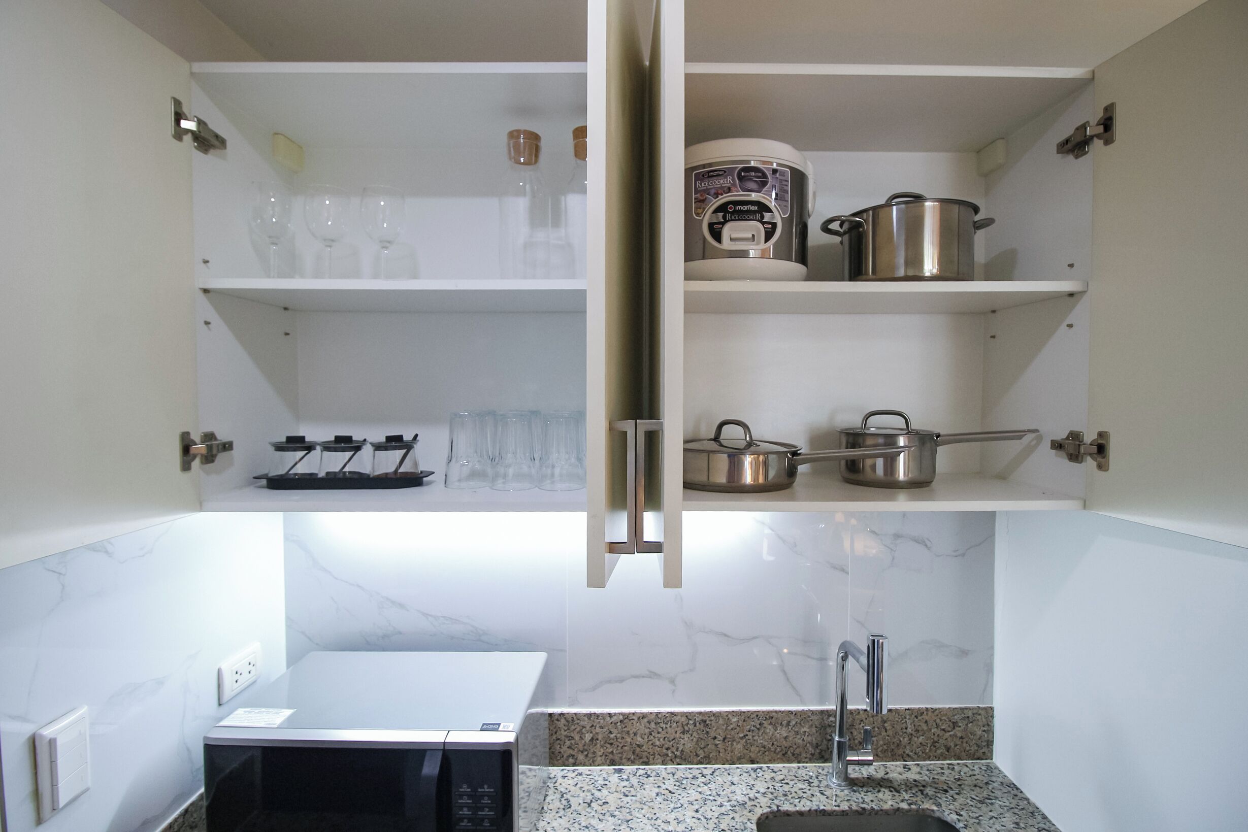 A kitchen with a microwave, granite countertop, and cabinets containing wine glasses, a coffee maker, and pots.
