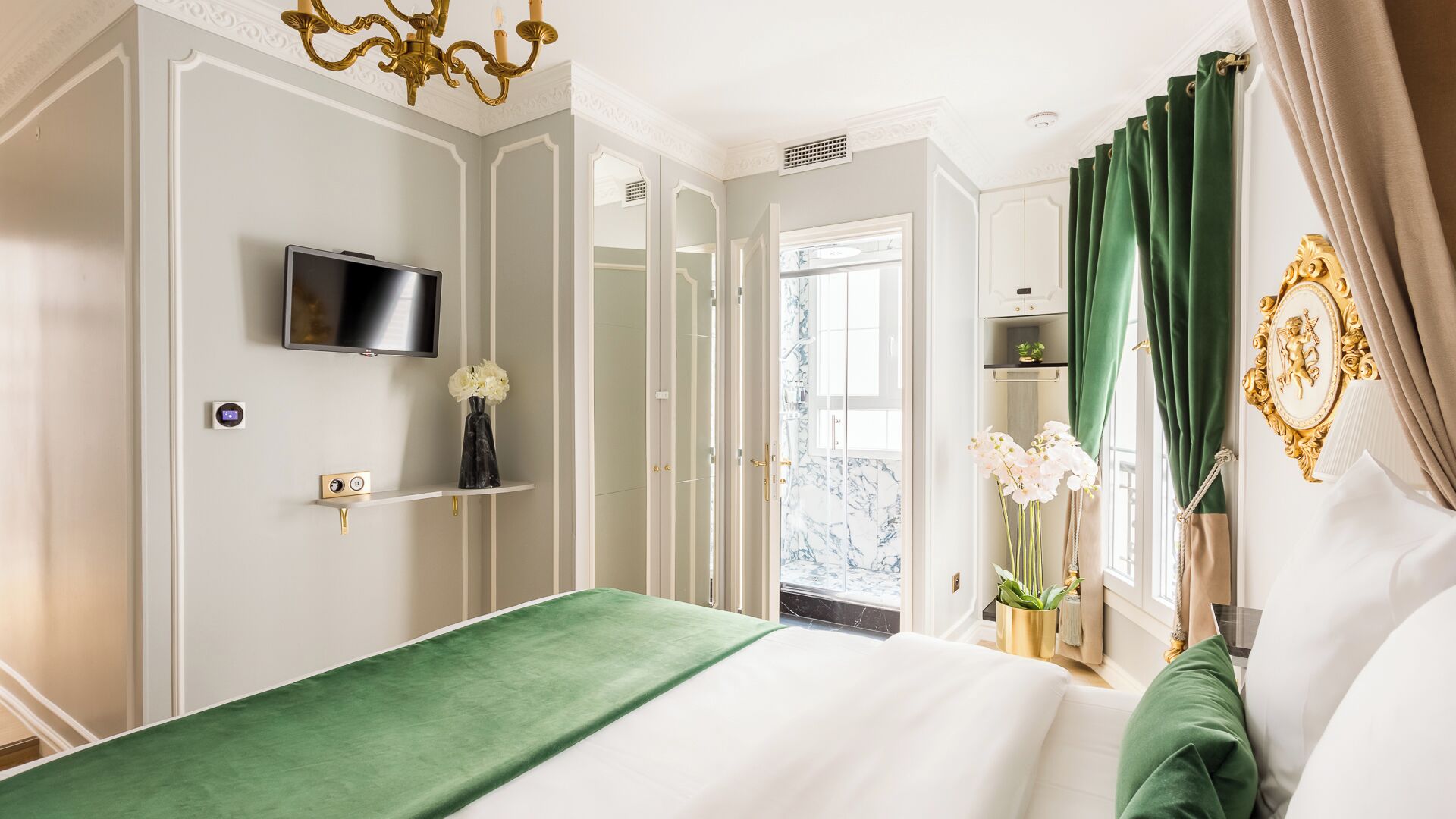 A bedroom with a bed, a television mounted on the wall, a small shelf with a vase, and a door leading to a balcony.