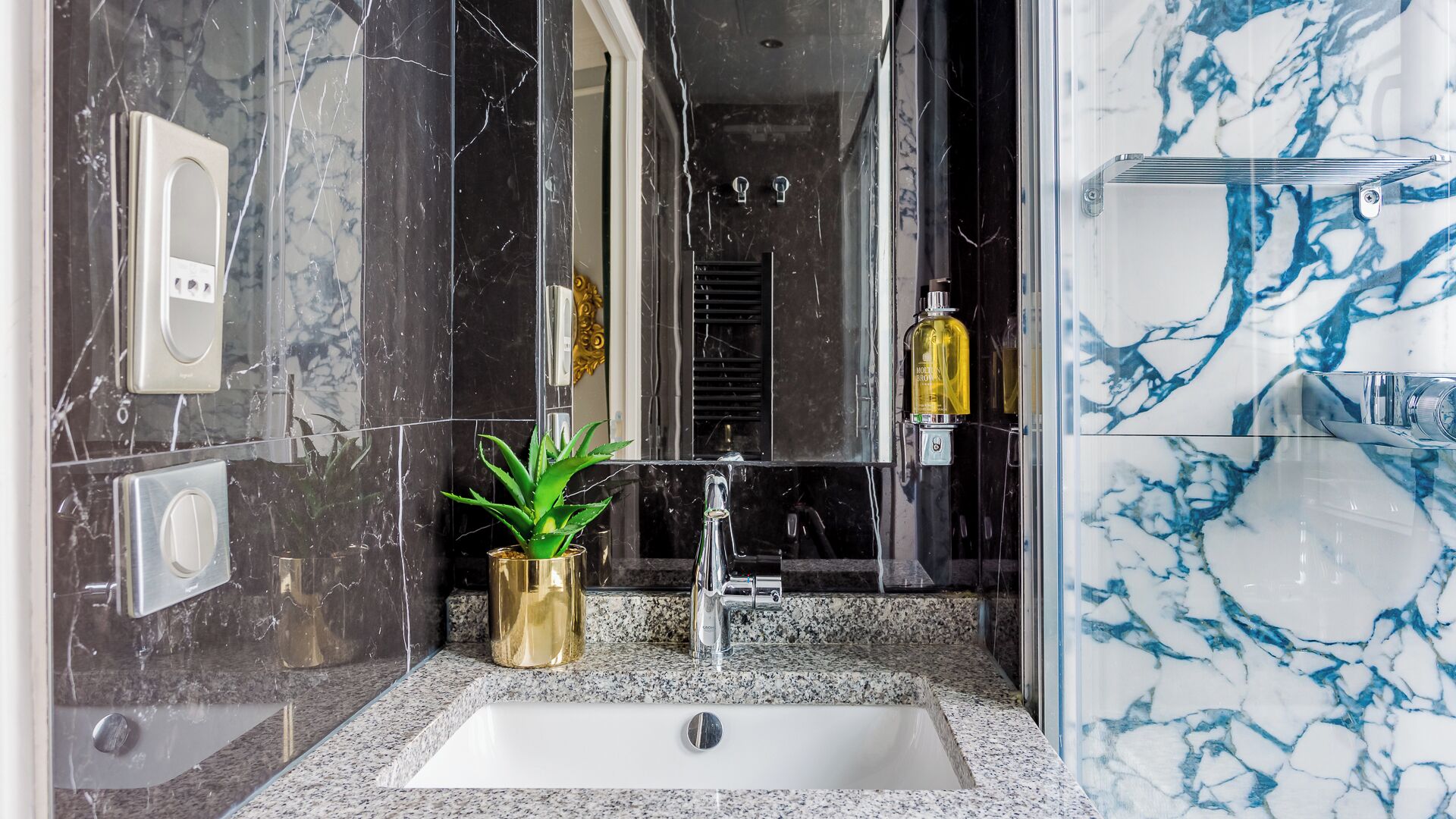 A bathroom with a marble sink, a mirror, a potted plant, and a bottle of lotion.
