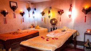 Une pièce équipée de deux tables de massage, chacune agrémentée de serviettes et de pétales de rose. On y trouve également des lampes fixées au mur et une petite table sur laquelle est posée une bougie.