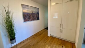 Un couloir avec un plancher en bois, une armoire blanche à plusieurs portes, une plante en pot et un tableau représentant un paysage urbain accroché au mur.