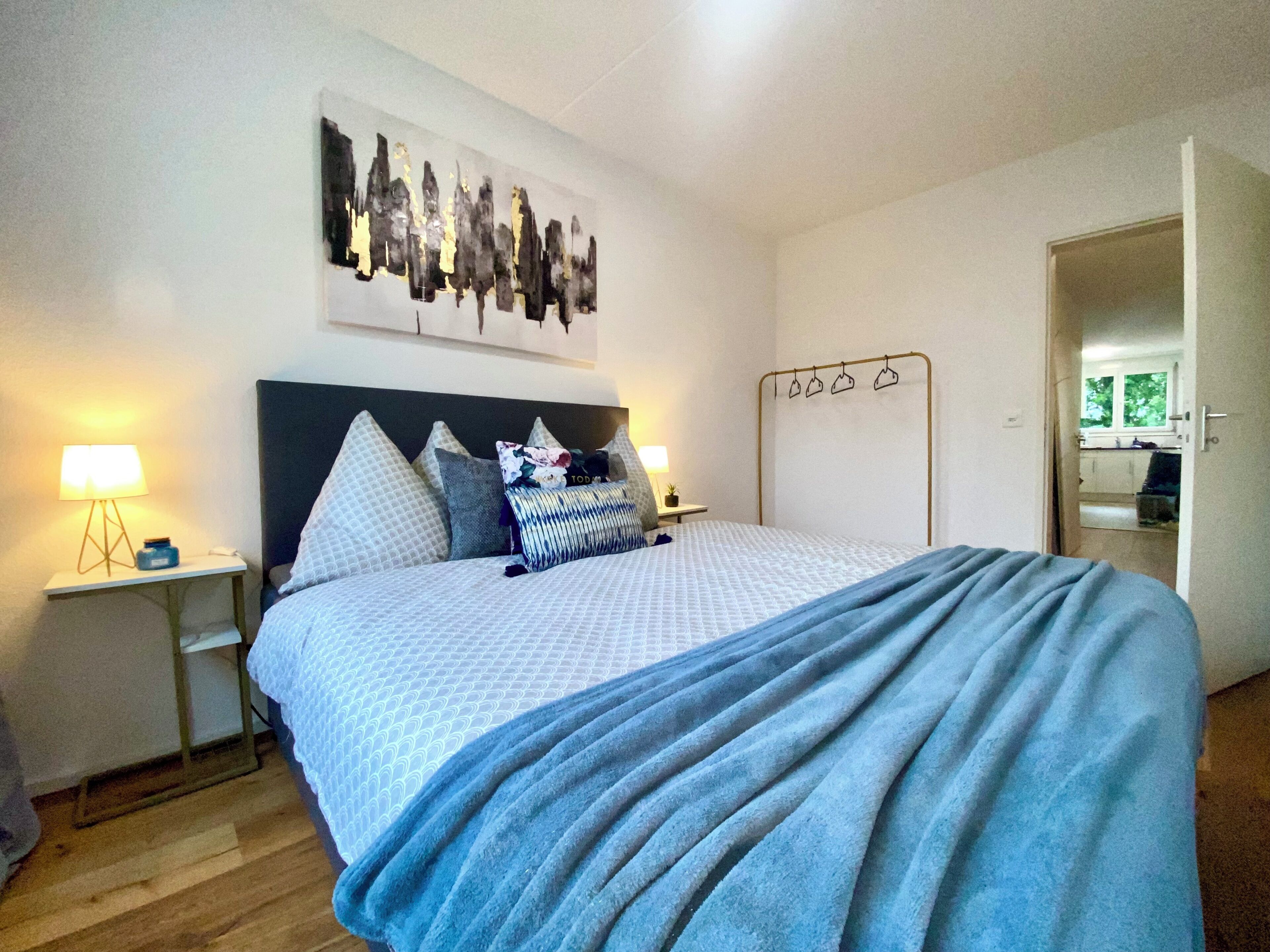 Une chambre à coucher bien rangée, avec un lit, deux tables de chevet, une œuvre d’art accrochée au mur et une porte donnant sur une salle de bain.
