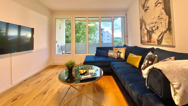 Un salon moderne avec un canapé bleu foncé, une table basse en verre et une grande fenêtre donnant sur des immeubles et des arbres.