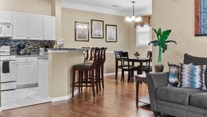 Una sala de estar con un sofá gris, una zona de comedor con una mesa y sillas, y una cocina con gabinetes blancos y salpicadero de azulejos.