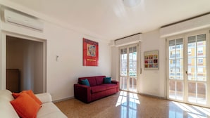 A spacious living room with a red sofa, a white sofa with orange cushions, a large window with a view of buildings, and an air conditioning unit. - Spacious flat near to Central Rome (Rome)