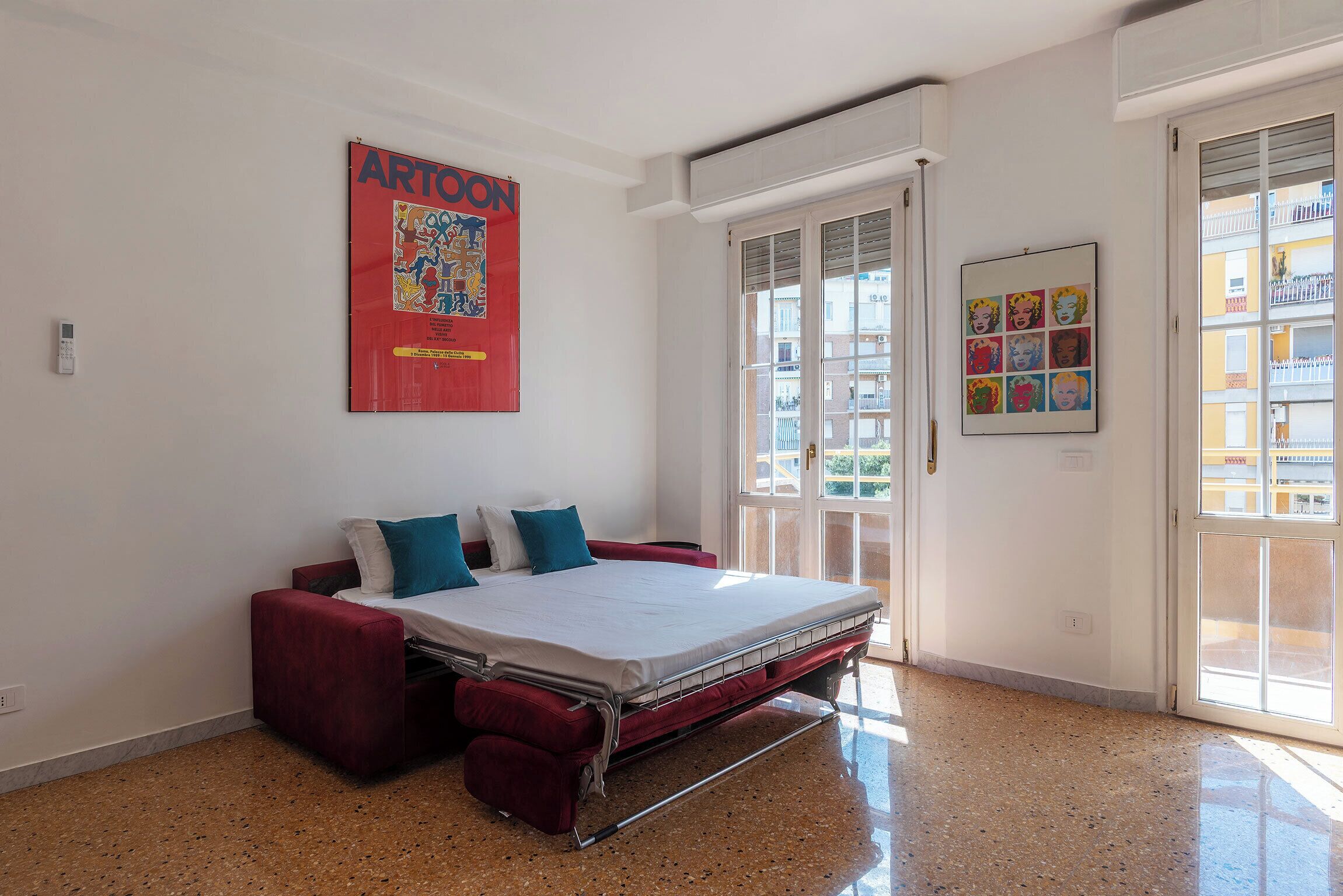 A bedroom with a bed, a red sofa, a window with a view of buildings, and a poster on the wall.