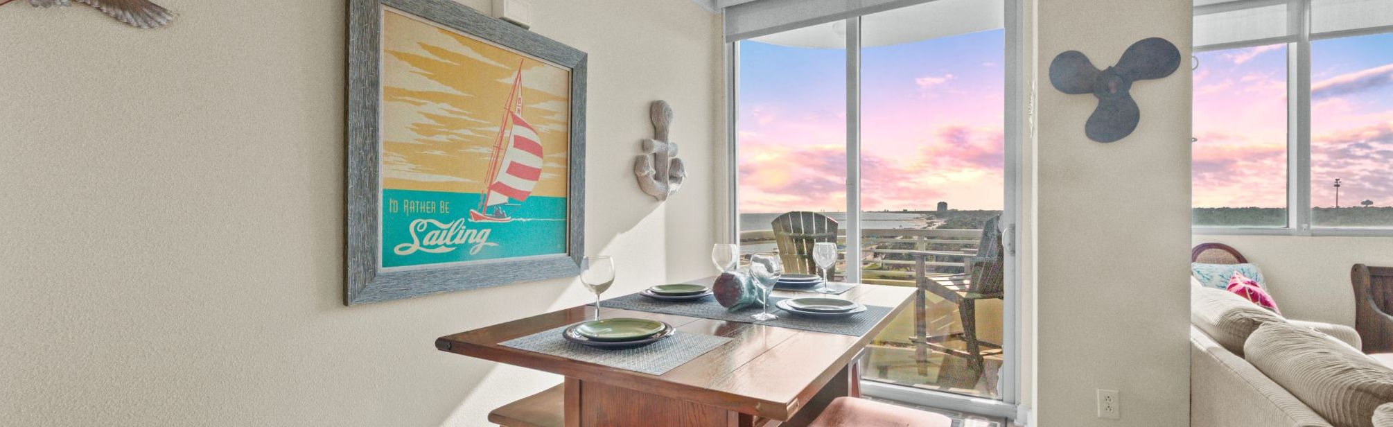 Un coin repas avec une table dressée pour quatre personnes, une grande fenêtre offrant une vue sur le coucher de soleil et une affiche encadrée accrochée au mur.