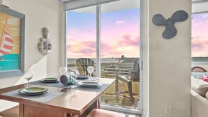 Een eetgedeelte met een tafel voor vier personen, een groot raam met uitzicht op een zonsondergang en een lijst met een poster aan de muur.