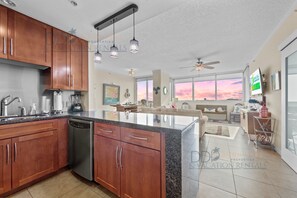 Een moderne keuken met houten kastjes, een vaatwasser en een groot eiland. Er is een zithoek met een bank en een televisie. De kamer heeft grote ramen met uitzicht op de zonsondergang.