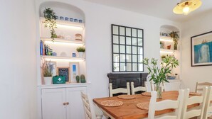 Un comedor con mesa de madera y sillas blancas, rodeado de estanterías con diversos objetos y una chimenea.