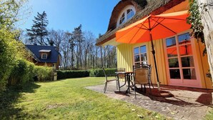 Une maison avec un toit de chaume et un parasol orange vif, une terrasse dallée avec deux chaises, et une pelouse bien entretenue.