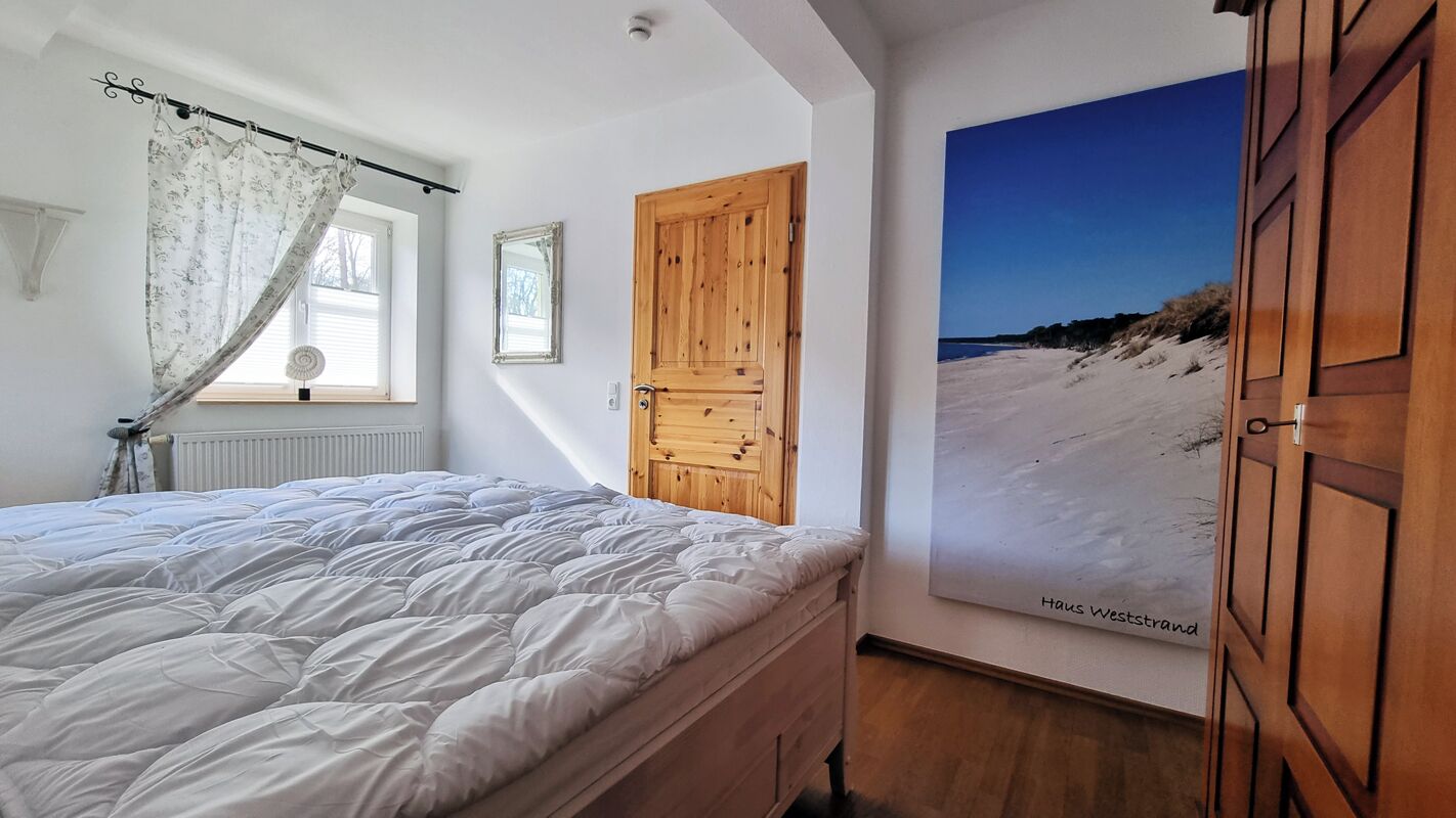 A bedroom with a large bed, a wooden wardrobe, a window with a curtain, and a beach-themed poster on the wall.