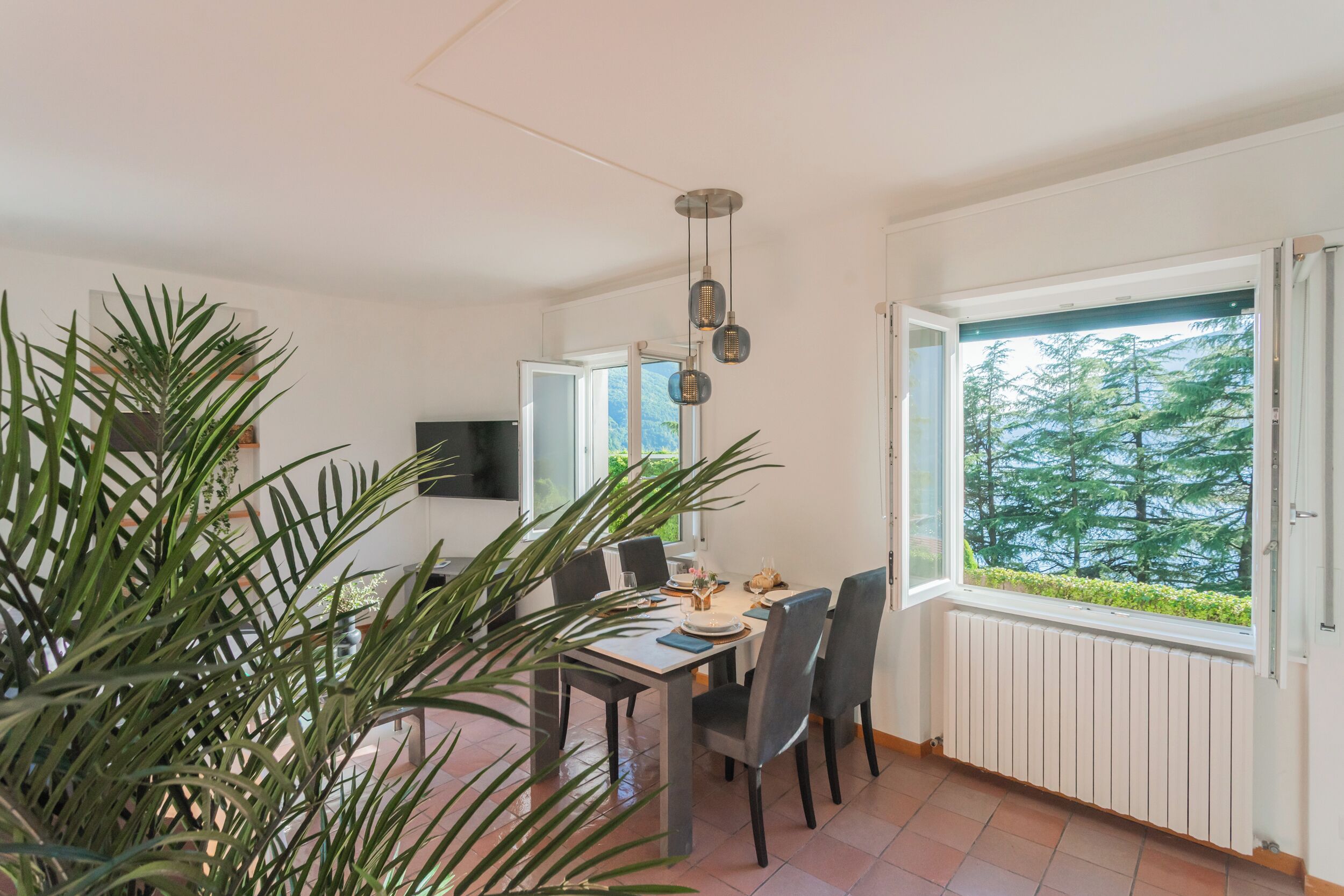 Een eetgedeelte met een tafel voor vier personen, een groot raam met uitzicht op bomen, en een potplant op de voorgrond.