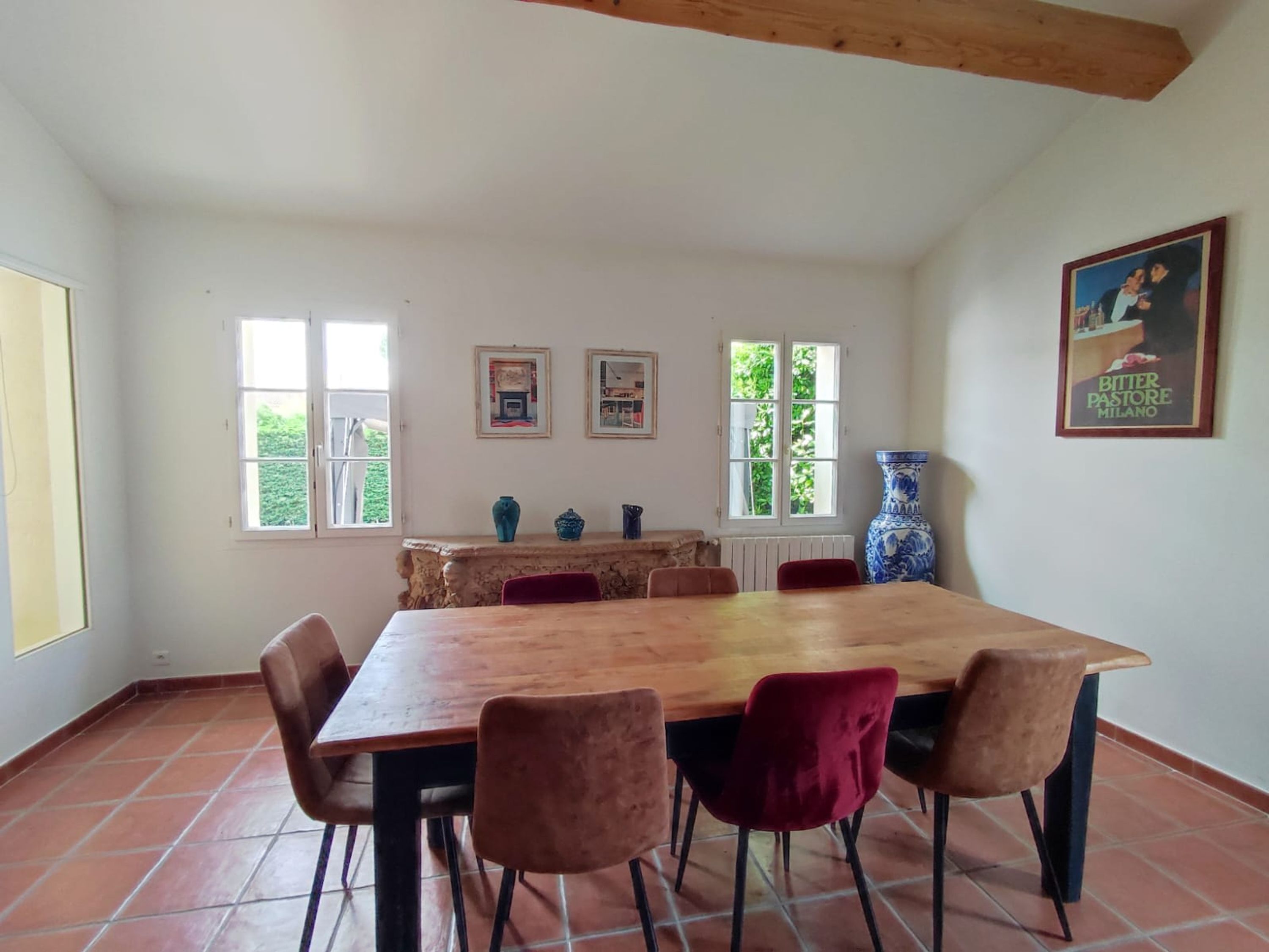 Un comedor con una mesa de madera y seis sillas, dos ventanas con vistas a zonas verdes, un jarrón azul y un cuadro en la pared.