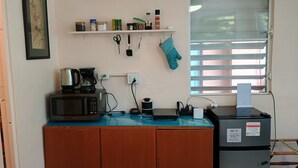 A kitchenette with a microwave, coffee maker, and various kitchen items on a counter. There is a window with blinds, a small shelf with spices, and a framed picture on the wall. - Room w/ba AC Wi-Fi in Esperanza Apt. 1 (Vieques)
