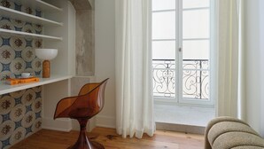 A room with a wooden floor, a white shelf with a bowl and books, a chair, and a window with white curtains and a balcony railing.