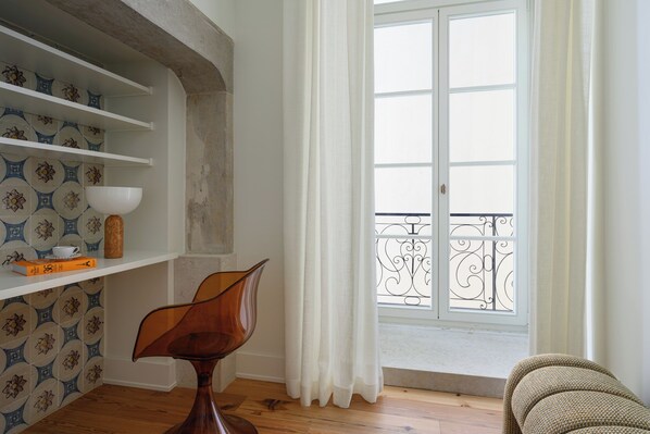Une pièce avec un plancher en bois, une étagère blanche sur laquelle se trouvent un bol et des livres, une chaise et une fenêtre avec des rideaux blancs et une balustrade de balcon.