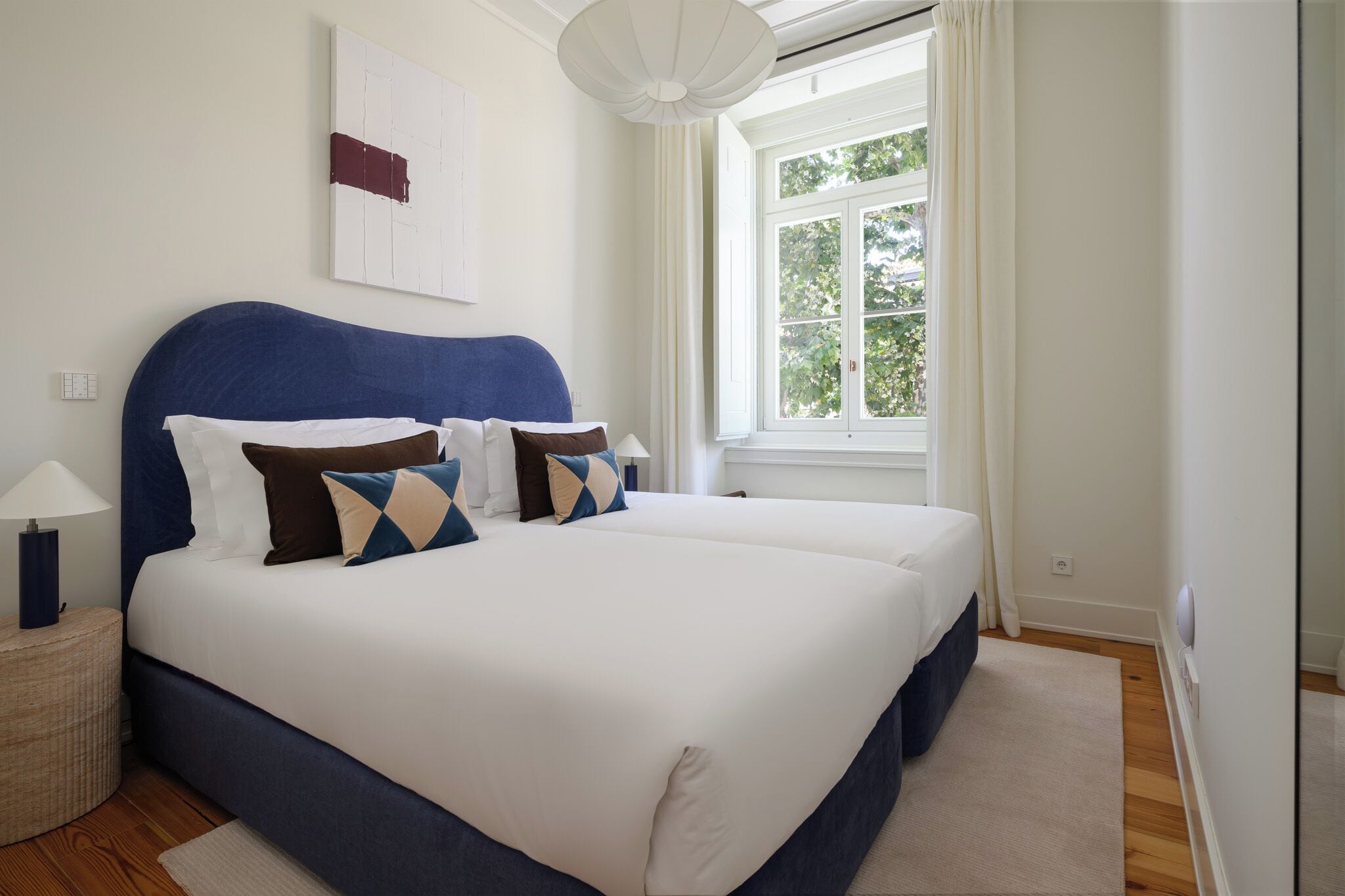 Cama doble con cabecera azul, ropa de cama blanca y almohadas estampadas. Hay una mesita de noche con una lámpara y una alfombra pequeña. Una ventana con cortinas blancas deja entrar luz natural.