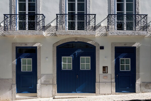 Un immeuble avec trois portes bleues, numérotées 81, 83 et 83a, et un balcon orné de balustrades en fer forgé.