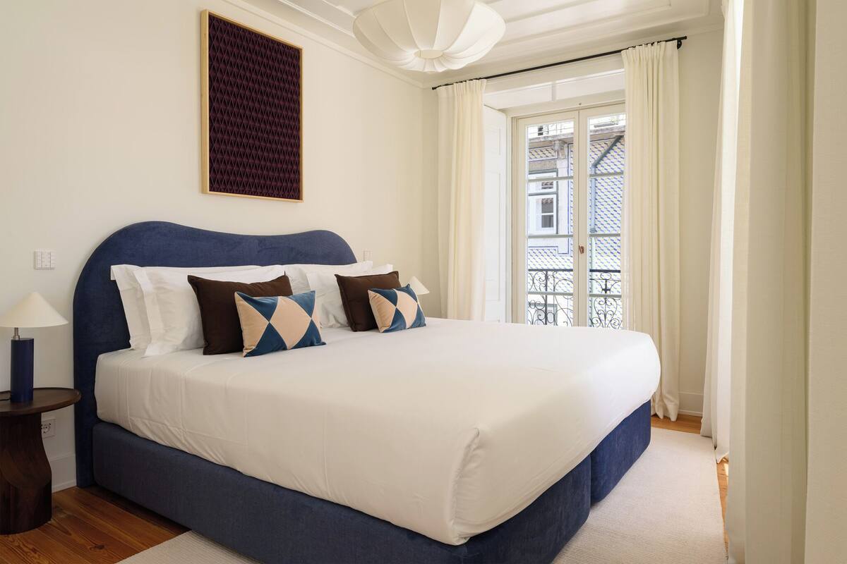 Een slaapkamer met een groot bed, een nachtkastje, een lamp, een raam met gordijnen en uitzicht op gebouwen.