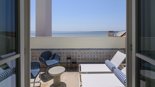 A balcony with a view of the sea, featuring a tiled wall, two blue patterned chairs, a small table, and a white lounge chair.