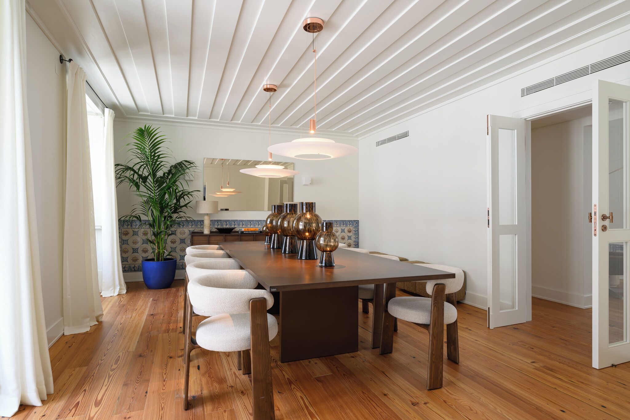 Een eethoek met een lange tafel, meerdere stoelen, een kamerplant en hanglampen.
