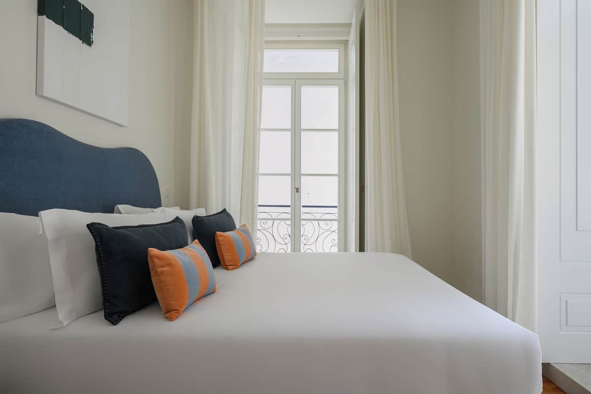 Een net opgemaakt bed met witte beddengoed en meerdere kussens, een groot raam met gordijnen en een witte deur.