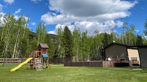 A wooden play structure with a slide, surrounded by a fenced yard and a building with a garage. - Cougar Mountain Lodge & Resort Ltd. (Valemount)