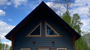 A cabin with a dark wooden exterior, a white door, and two windows on the front. There is a sign that reads "CABIN 9" and a blue recycling bin. - Cougar Mountain Lodge & Resort Ltd. (Valemount)
