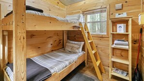 A wooden cabin room with bunk beds, a ladder, a window, and a shelf with items.