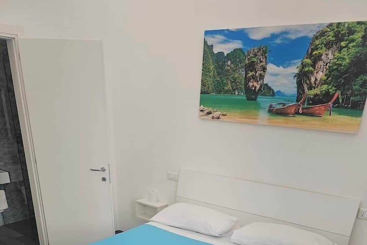 Une chambre à coucher avec un lit, des murs blancs, une porte et un tableau représentant une scène de plage.