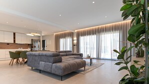 Een moderne woonkamer met een grijze hoekbank, een eethoek met een kleine tafel en stoelen, en grote ramen met gordijnen.
