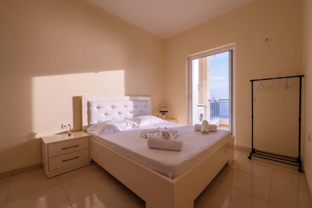 A bedroom with a bed, a bedside table, a nightstand, a window, and a balcony.