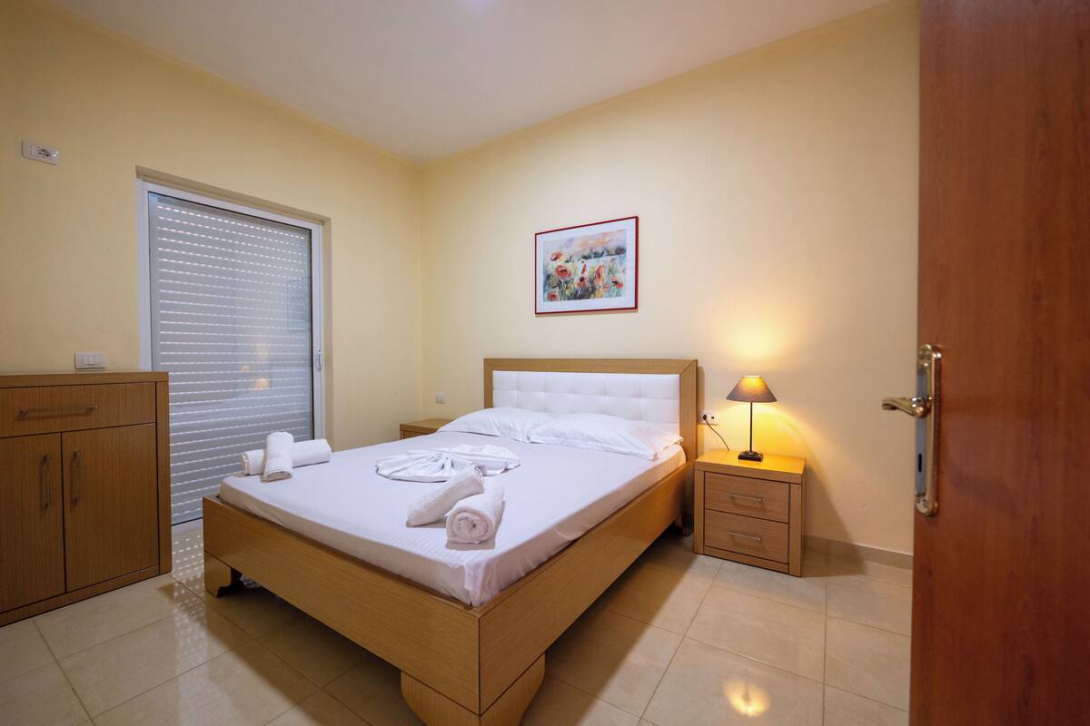 A neatly made bed with white linens and towels, a bedside lamp, a wooden nightstand, a wooden wardrobe, and a window with blinds.