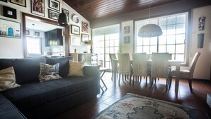 A living room with a dark-colored sofa, a dining table with chairs, and a large window with blinds.