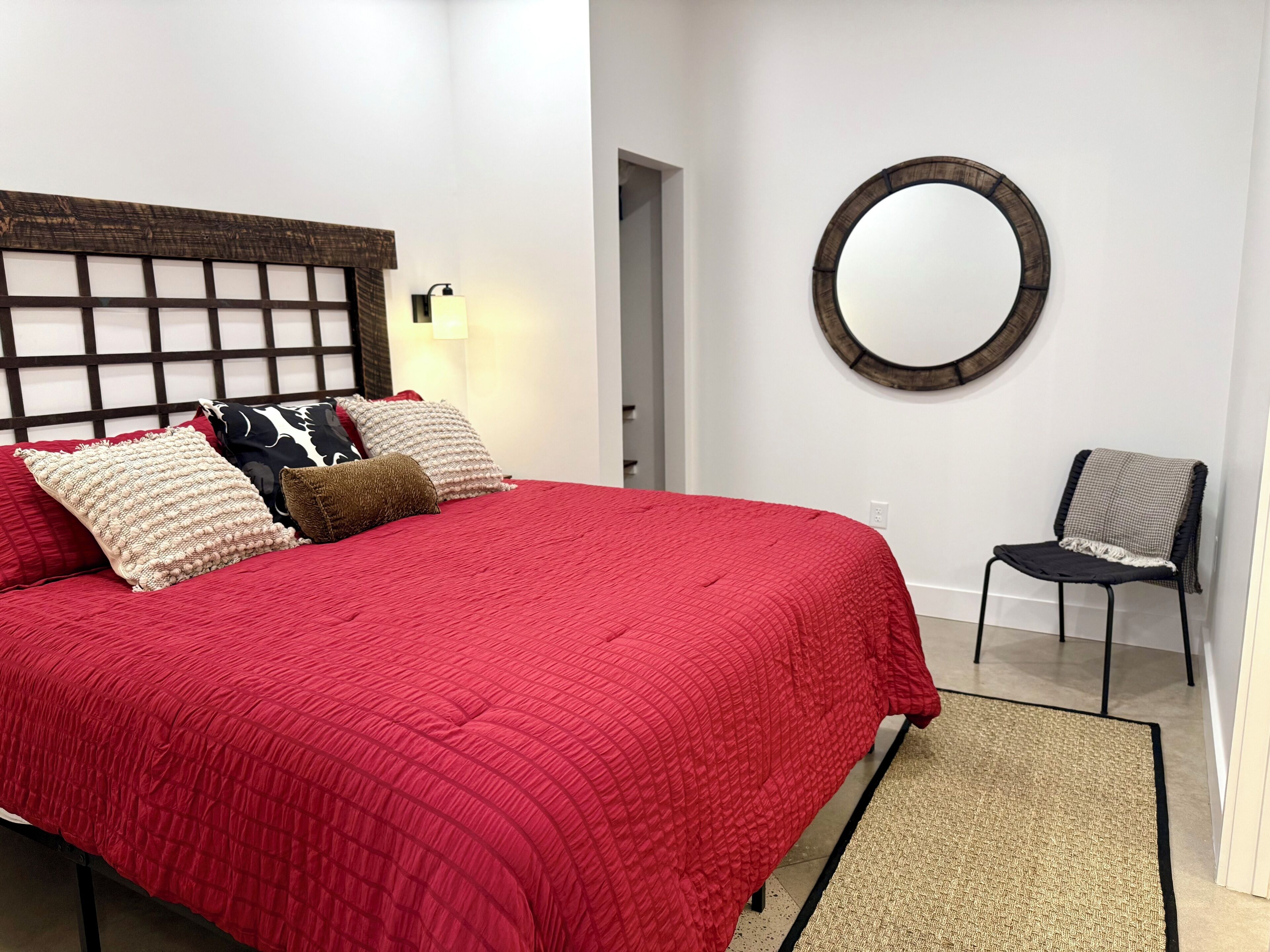 A bedroom with a red bedspread, a wooden headboard, and a round mirror on the wall.