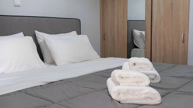 A bedroom with a bed, pillows, a grey quilted mattress, and a stack of white towels. - Petralona Antistratou Athens (Athens)