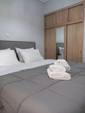 A bedroom with a bed, pillows, a grey quilted mattress, and a stack of white towels.