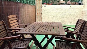A wooden table and chairs set on artificial grass under a covered area with a bamboo fence and green foliage backdrop. - Petralona Antistratou Athens (Athens)