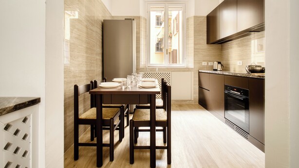 A modern kitchen with a dining area, featuring a built-in oven, a window with a view, and a light wooden floor.