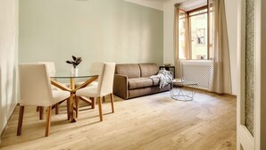 A modern living room with a glass dining table, two chairs, a sofa, and a window with curtains.