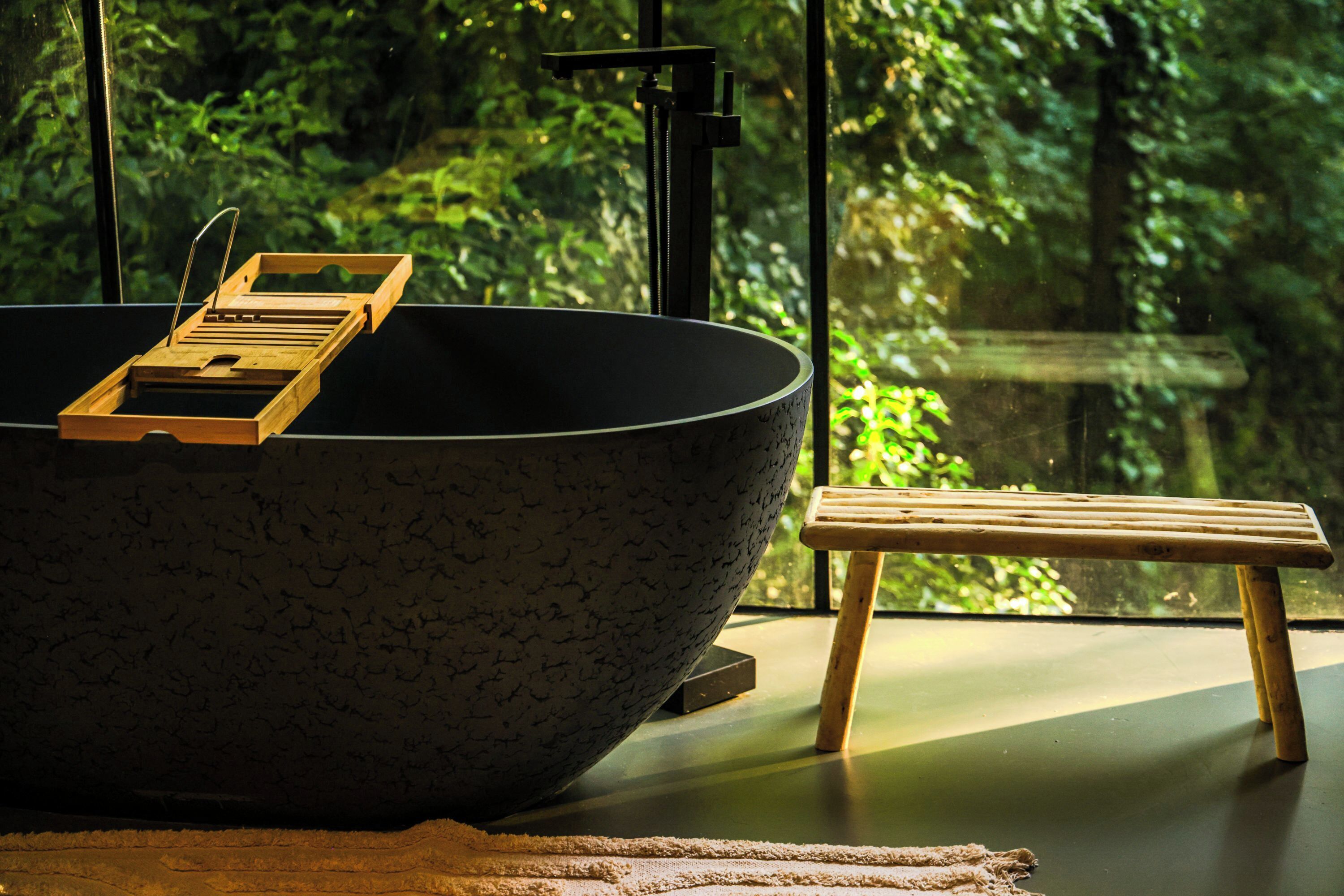 Un grand saladier noir, posé sur un plateau en bois, se trouve sur une table, à côté d’un tabouret en bois. Une fenêtre offre une vue sur la verdure.
