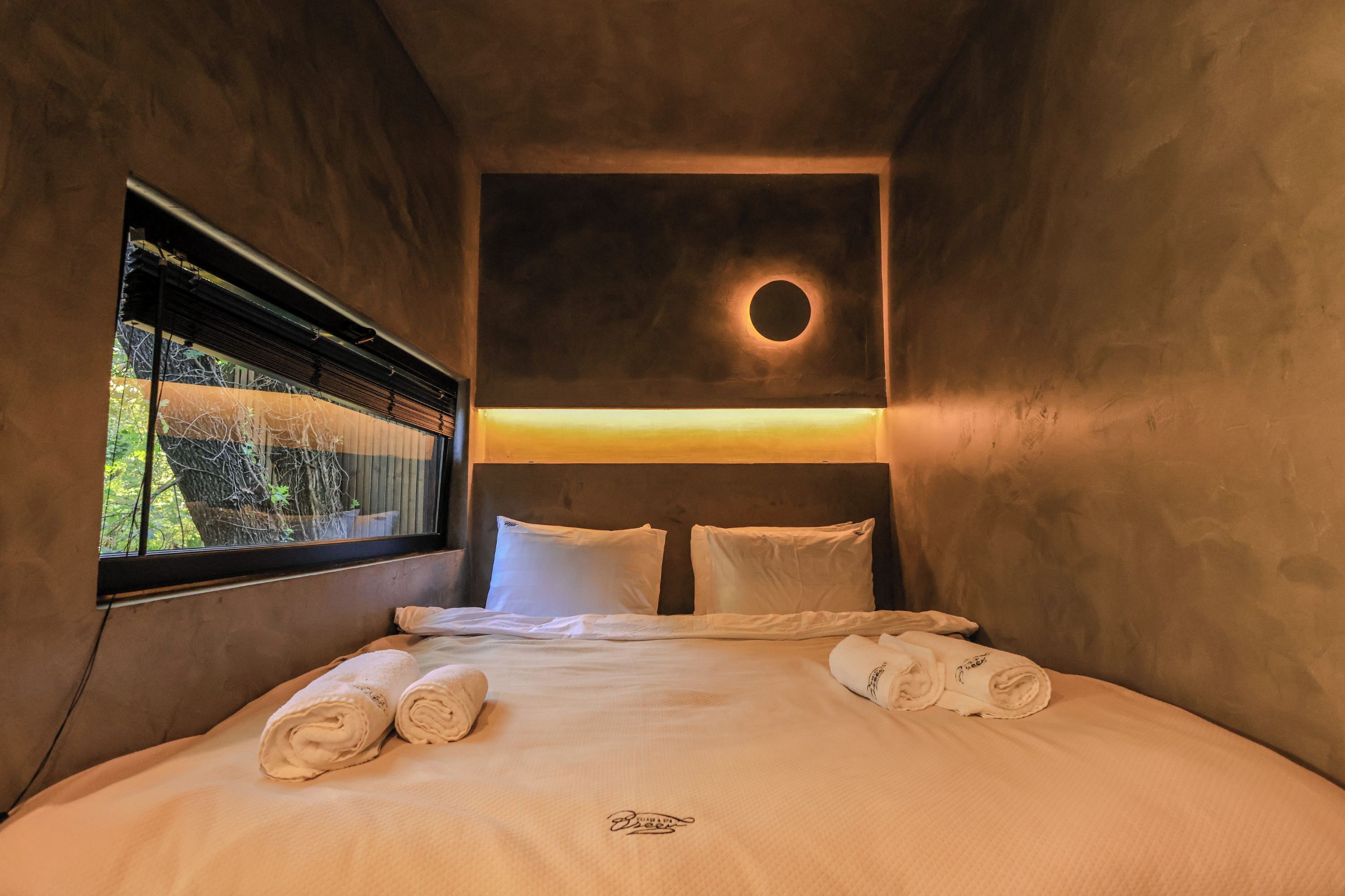 A cozy room with a bed, two pillows, a circular light fixture, and a window with blinds.
