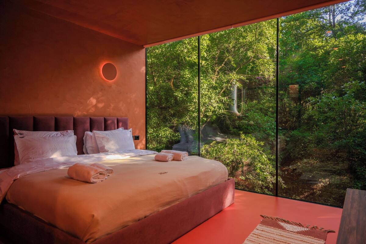 Une chambre moderne avec un grand lit, un luminaire circulaire et une vue sur une forêt grâce à de grandes fenêtres du sol au plafond.